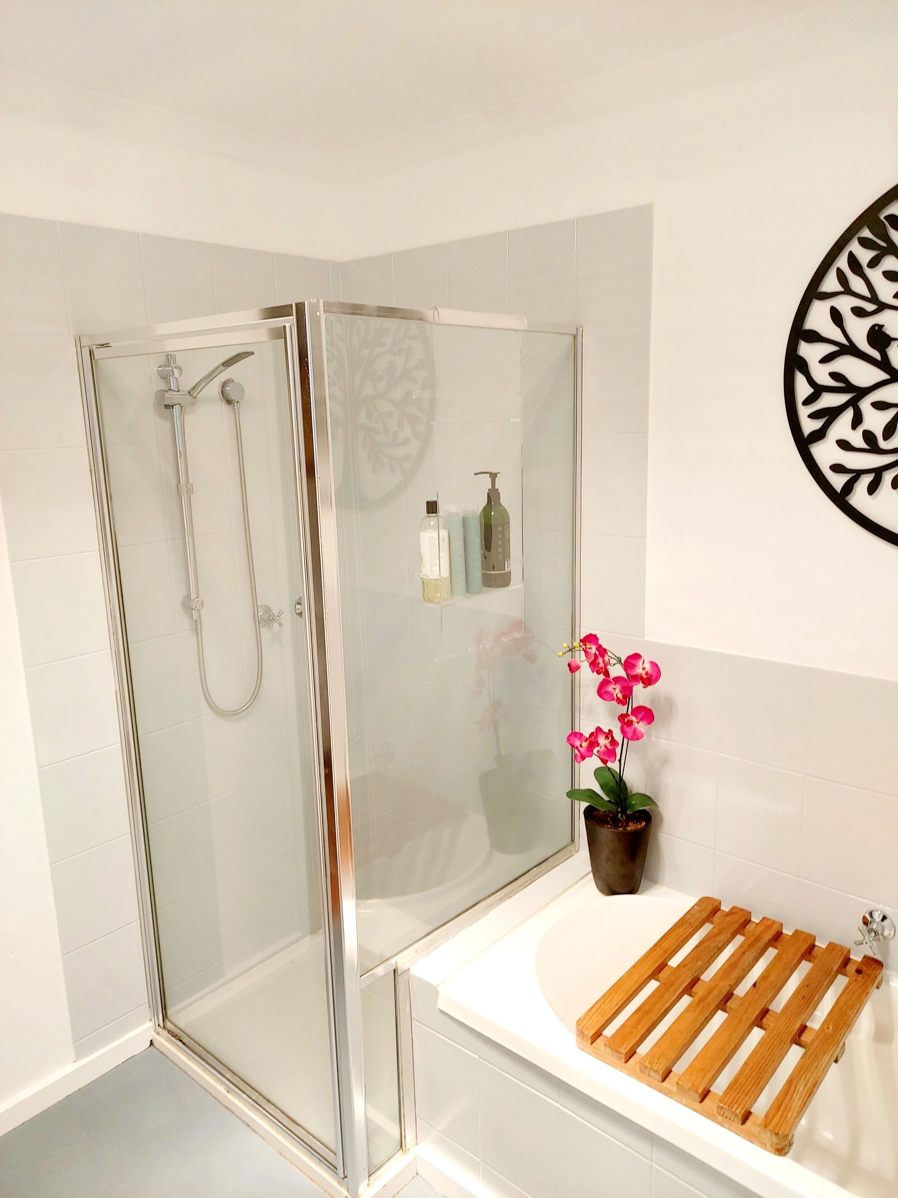 Une douche avec une porte en verre, un tapis de bain en bois, une orchidée en pot et un élément décoratif mural.