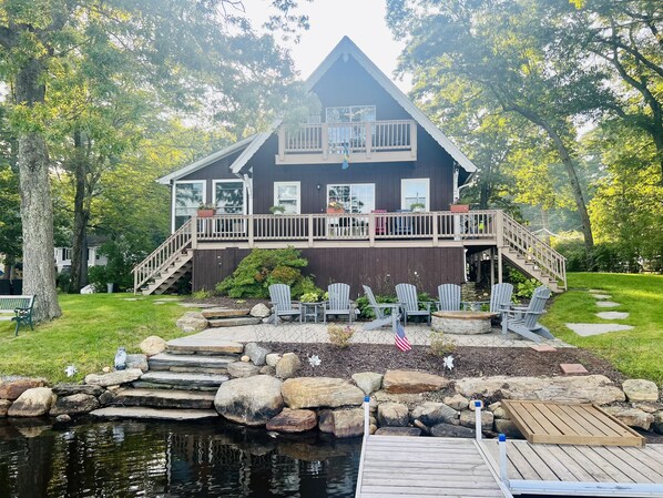 A two-story house with a deck, outdoor seating, and a pond. - Direct waterfront Lake House, stunning views Sleeps 8 on a private PENINSULA! (Sturbridge)