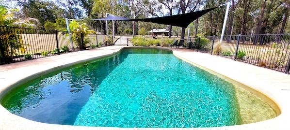 A rectangular swimming pool with clear blue water, surrounded by a black metal fence and a shaded area.