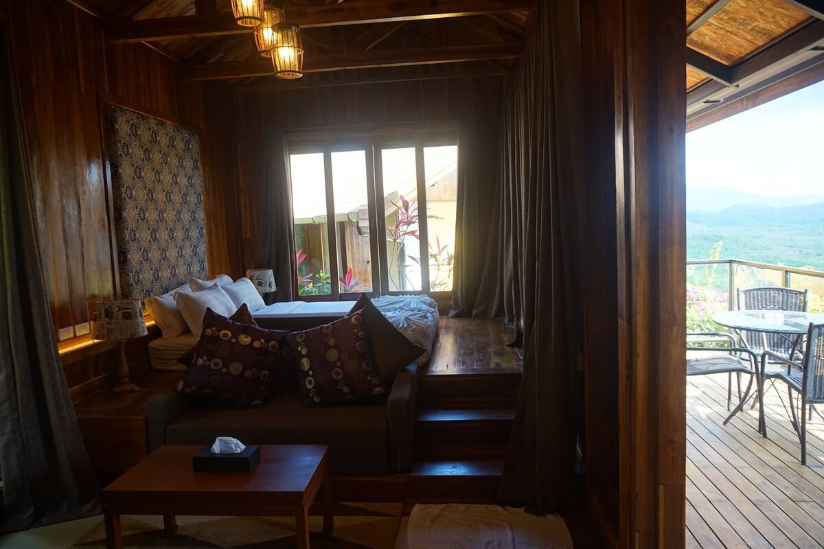 A cozy room with a bed, a sofa, and a table. There is a window with curtains and a view of greenery outside.