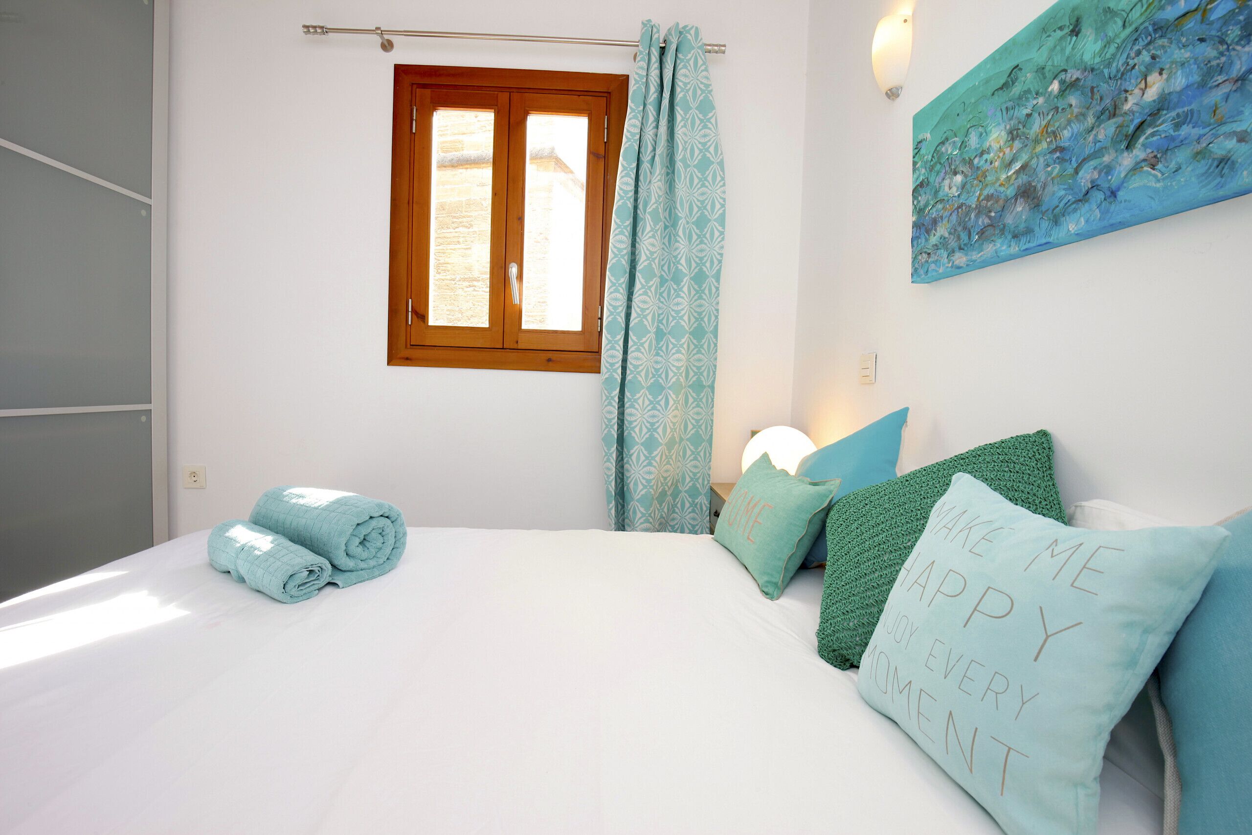 A bedroom with a bed, a window with curtains, a painting on the wall, and a pillow with a message.