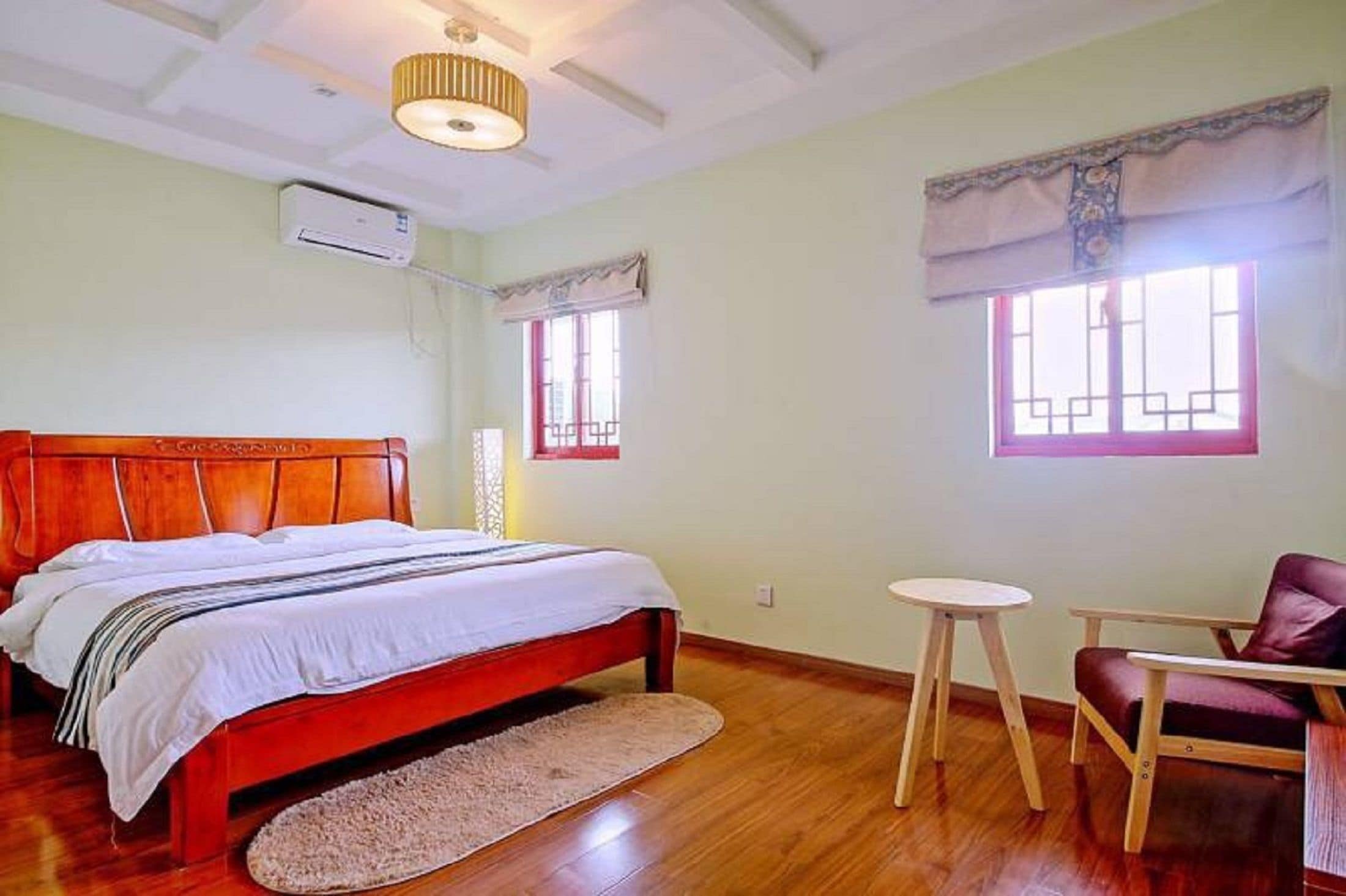 Ein Schlafzimmer mit einem hölzernen Bett, einem kleinen runden Tisch, einem Stuhl und zwei mit Vorhängen versehenen Fenstern.
