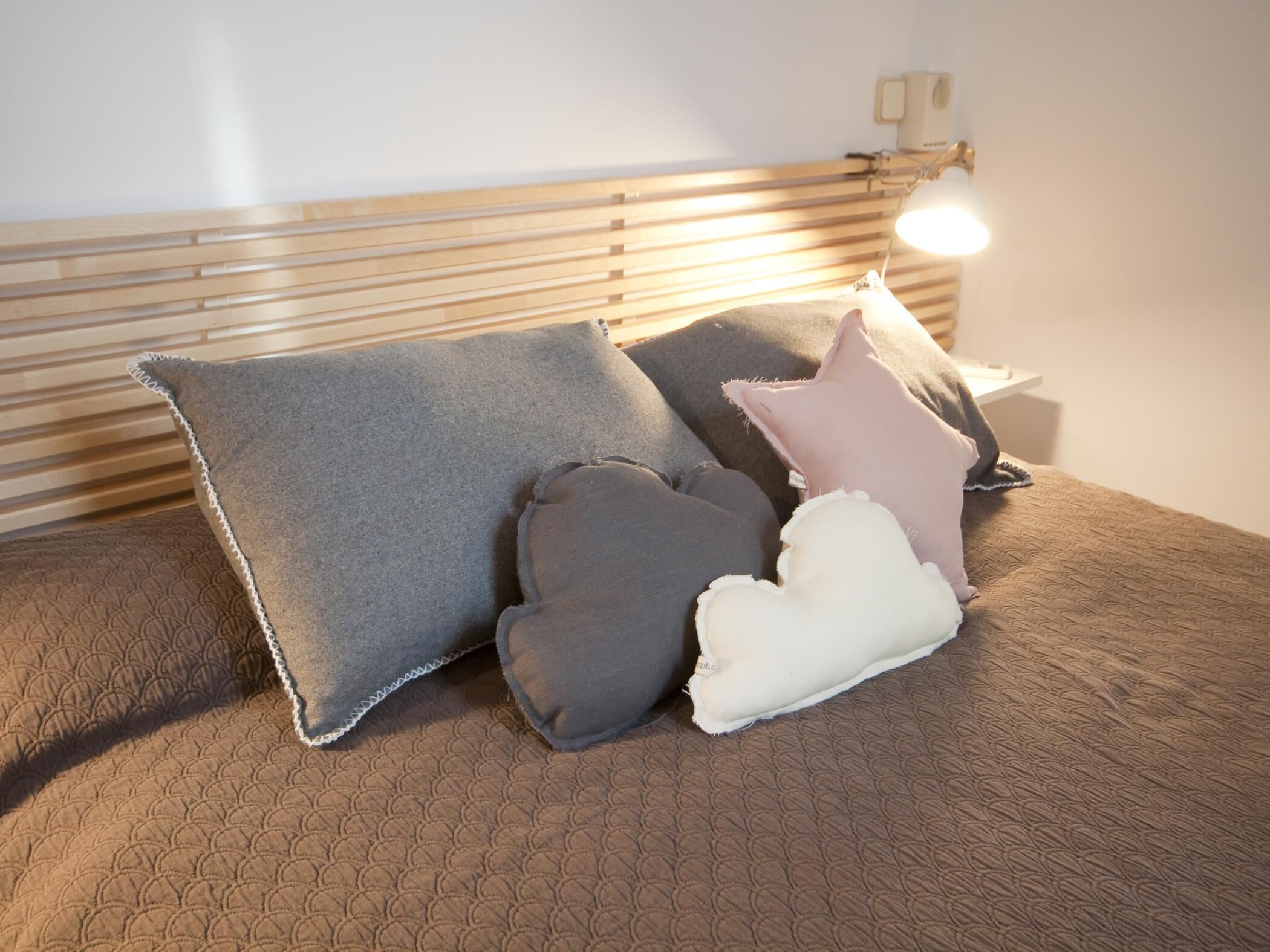 A bed with a textured brown cover, accompanied by three pillows in varying shades of gray and white.