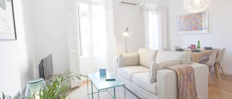 A bright living room with a white sofa, a small table, and a dining area with a table and chairs.