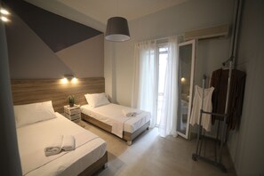 Une chambre d’hôtel avec deux lits, une petite table de chevet agrémentée d’une plante, un luminaire suspendu, une porte-fenêtre coulissante donnant sur la salle de bain et un porte-manteau.