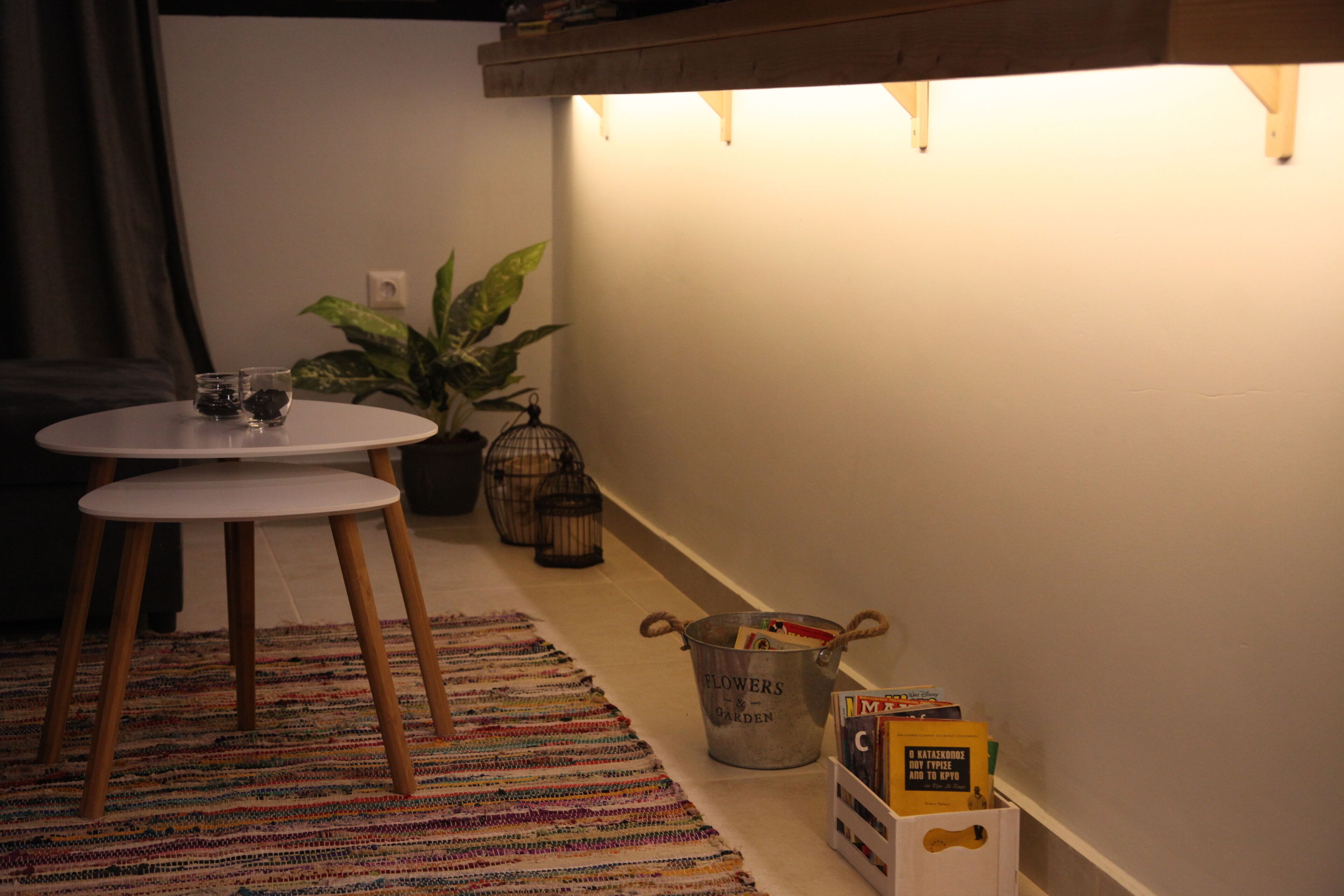 A cozy room with a round table, a patterned rug, a potted plant, and a metal bucket labeled "FLOWERS & GARDEN" on the floor.