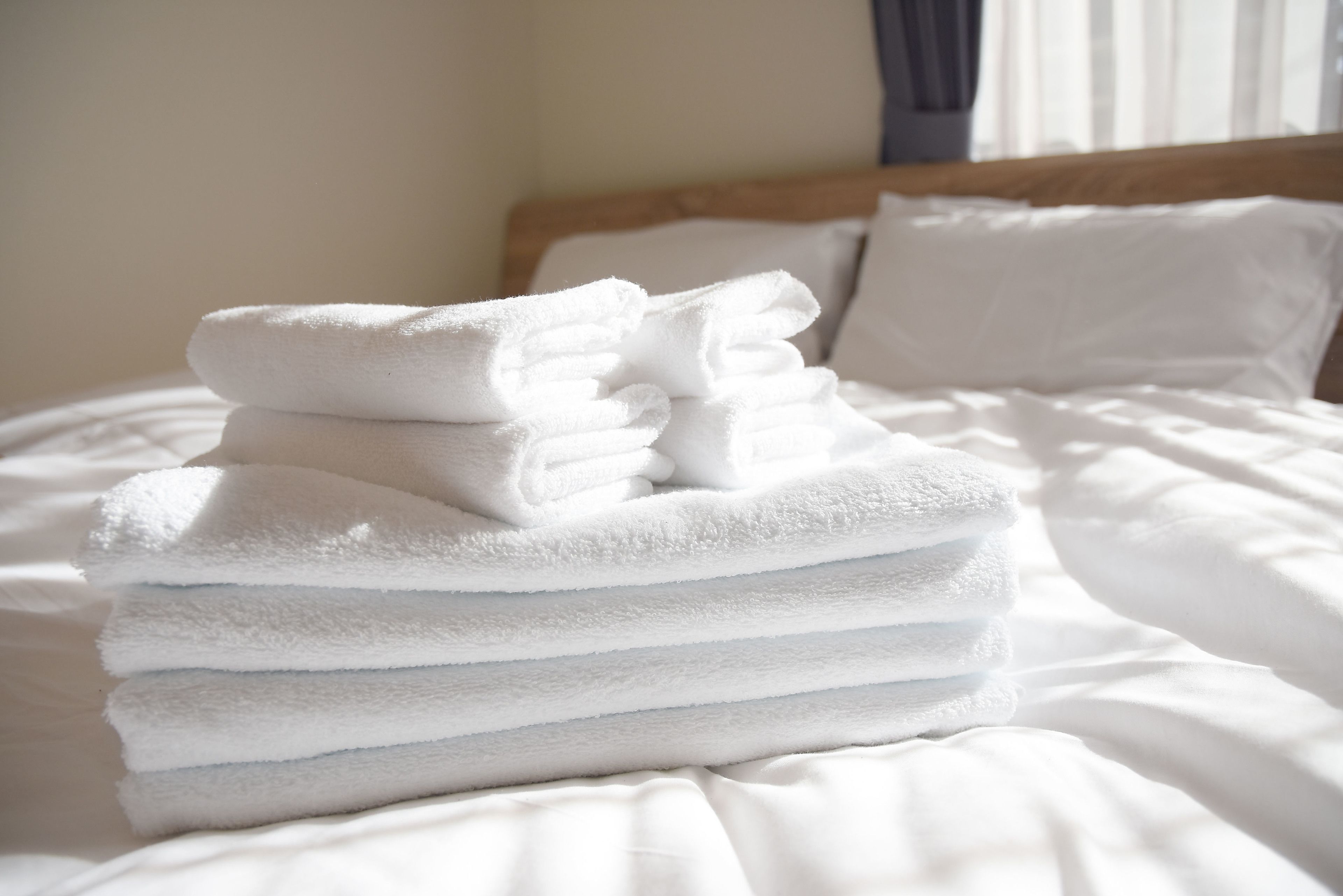A neatly stacked pile of white towels on a bed with white bedding and a wooden headboard.