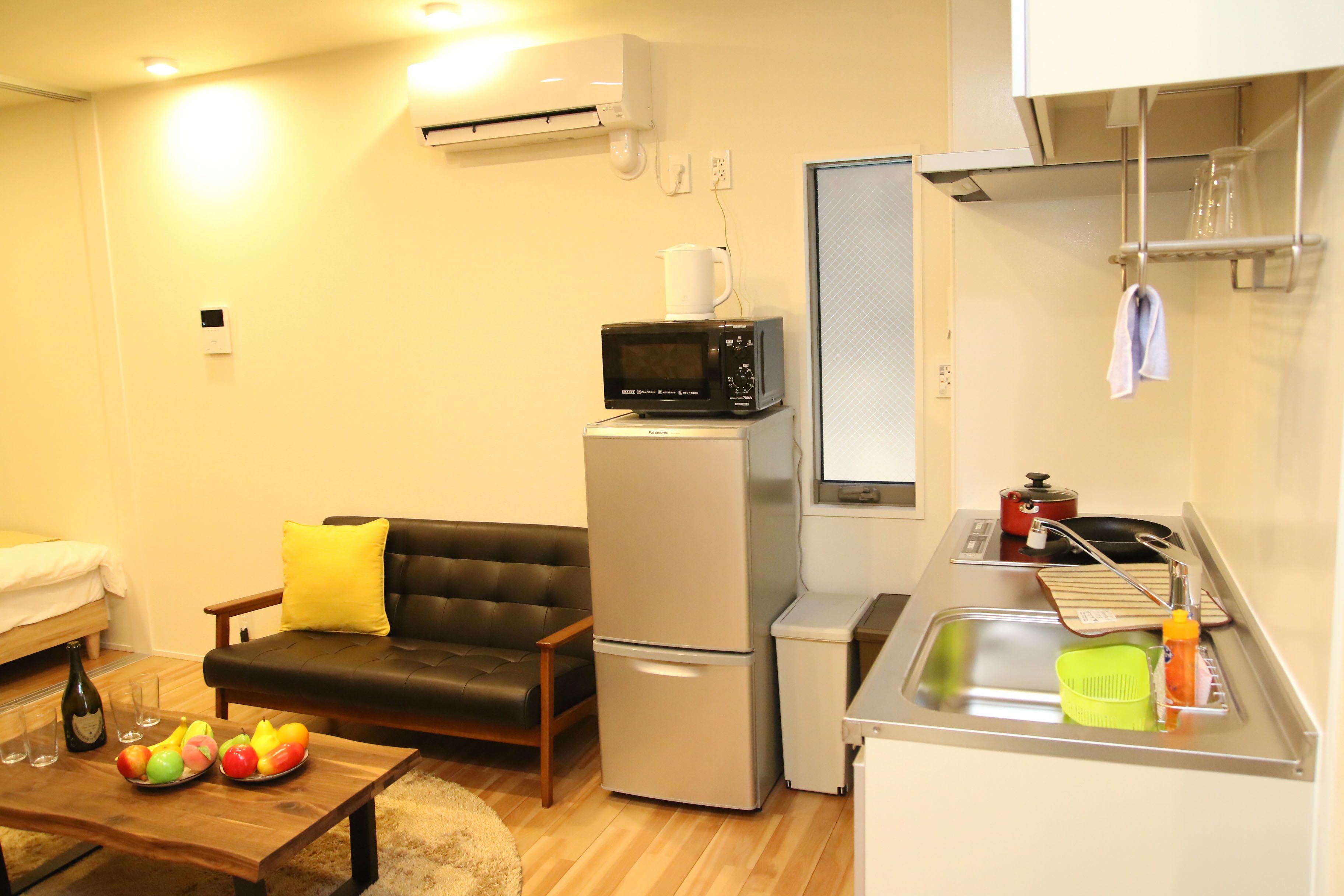 A compact living space with a sofa, a small kitchen area including a microwave and refrigerator, and a dining table with fruit.