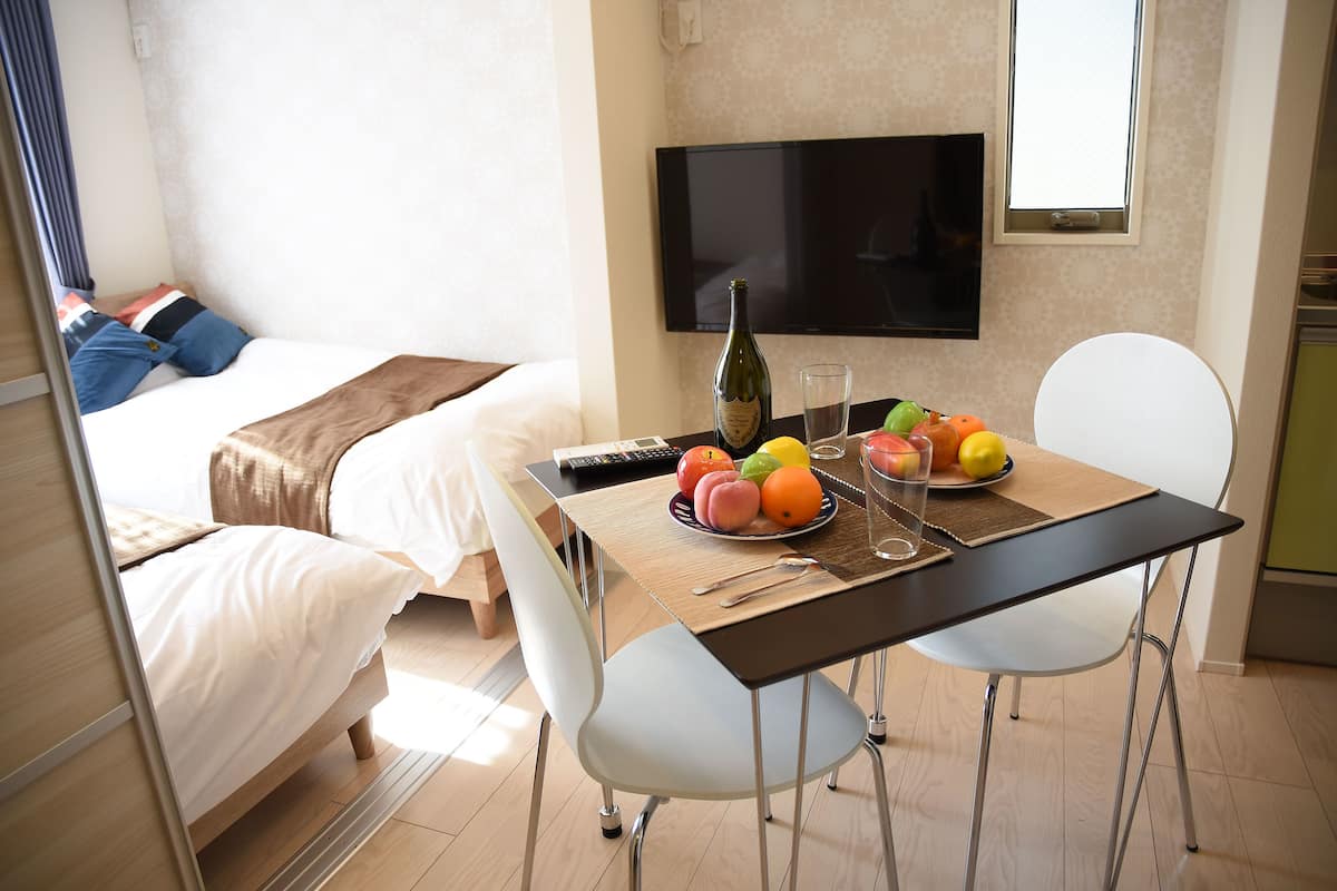 Une chambre d’hôtel avec un lit, un téléviseur fixé au mur, une table à manger avec des fruits et une bouteille, et une chaise.