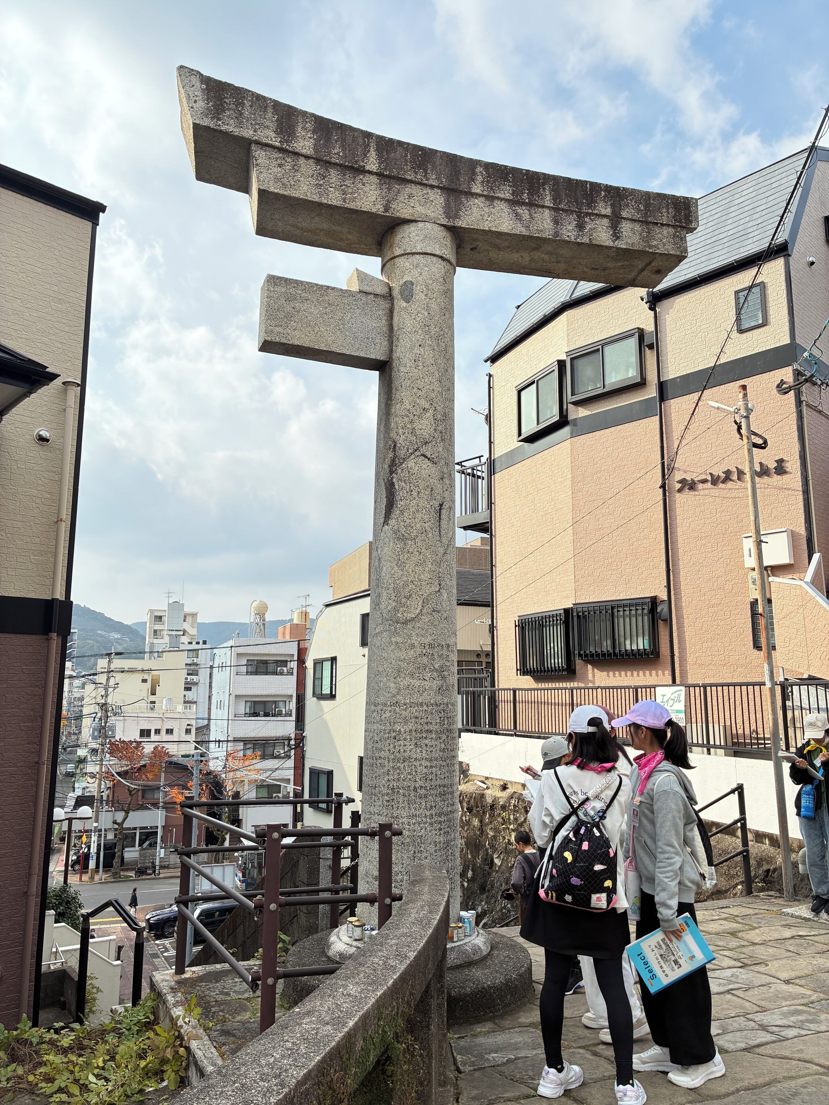 A stone monument with a cross-like structure, surrounded by residential buildings and a street.