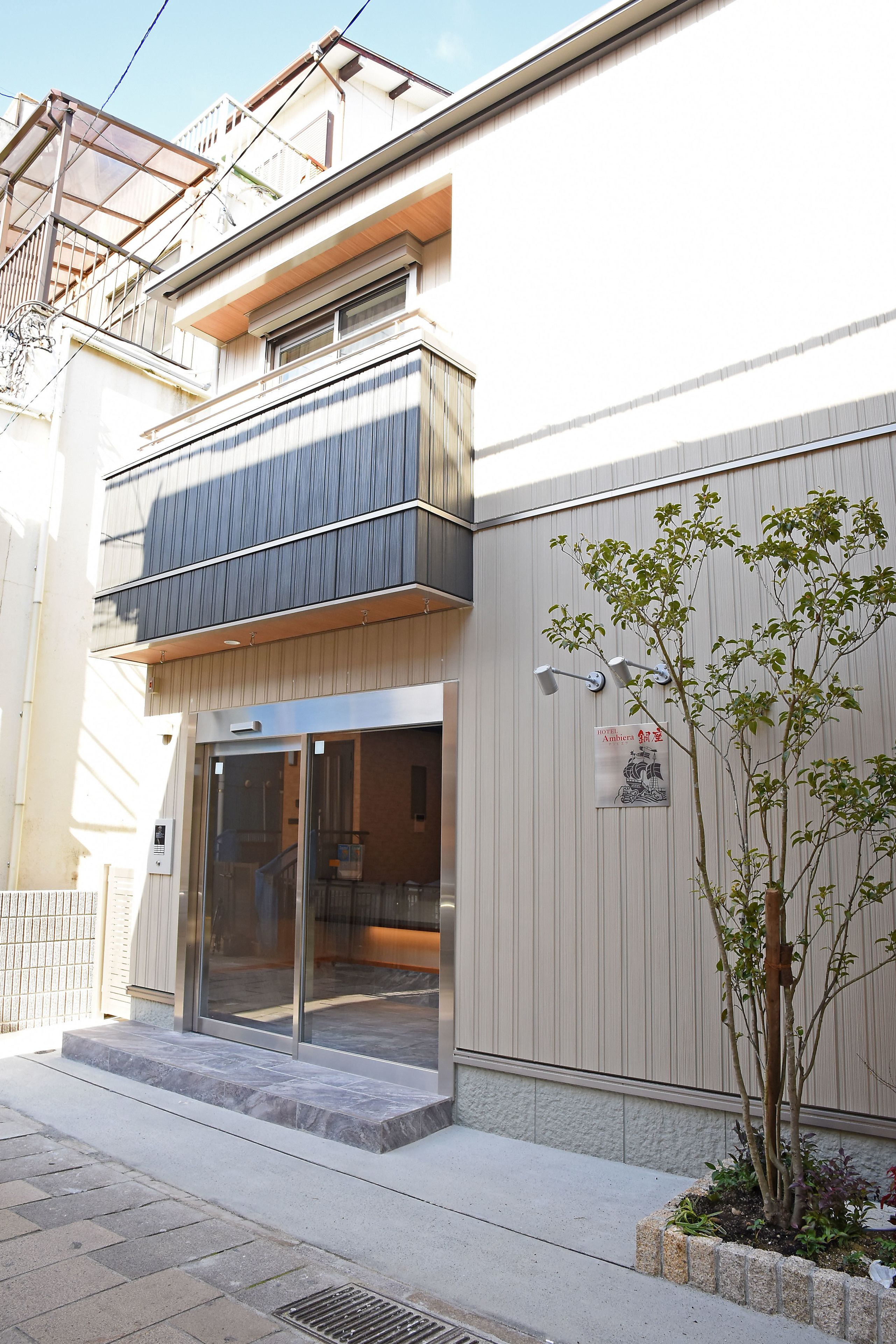 A modern building with a glass entrance, a small tree in the foreground, and a sign on the wall.