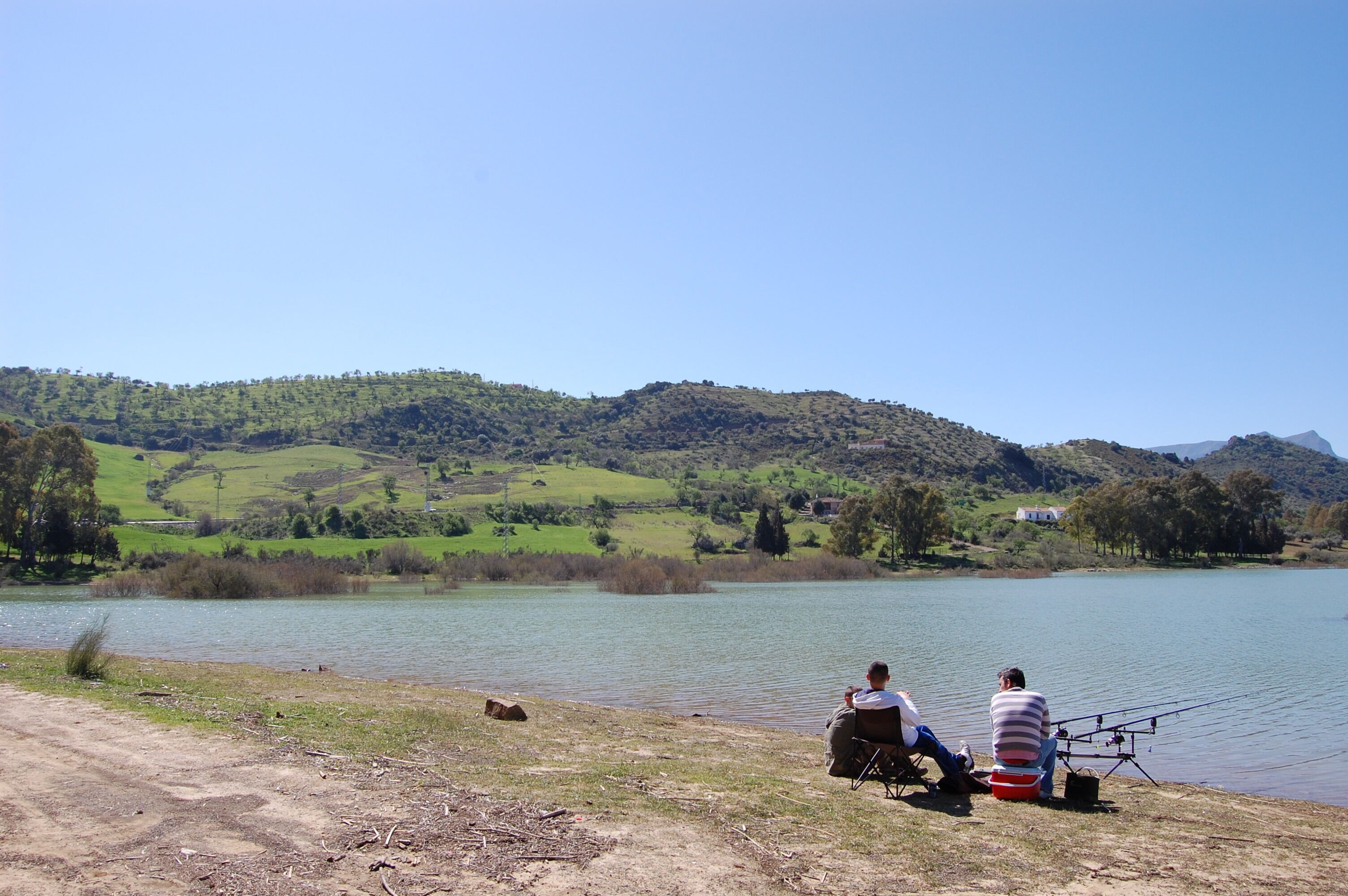 Zwei Personen sitzen an einem See mit Blick auf grüne Hügel im Hintergrund.