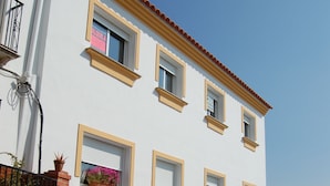 Un edificio de dos pisos con fachada blanca, marcos de ventanas amarillos y techo de tejas rojas. Cuenta con dos puertas de garaje y un pequeño balcón con una planta en maceta.