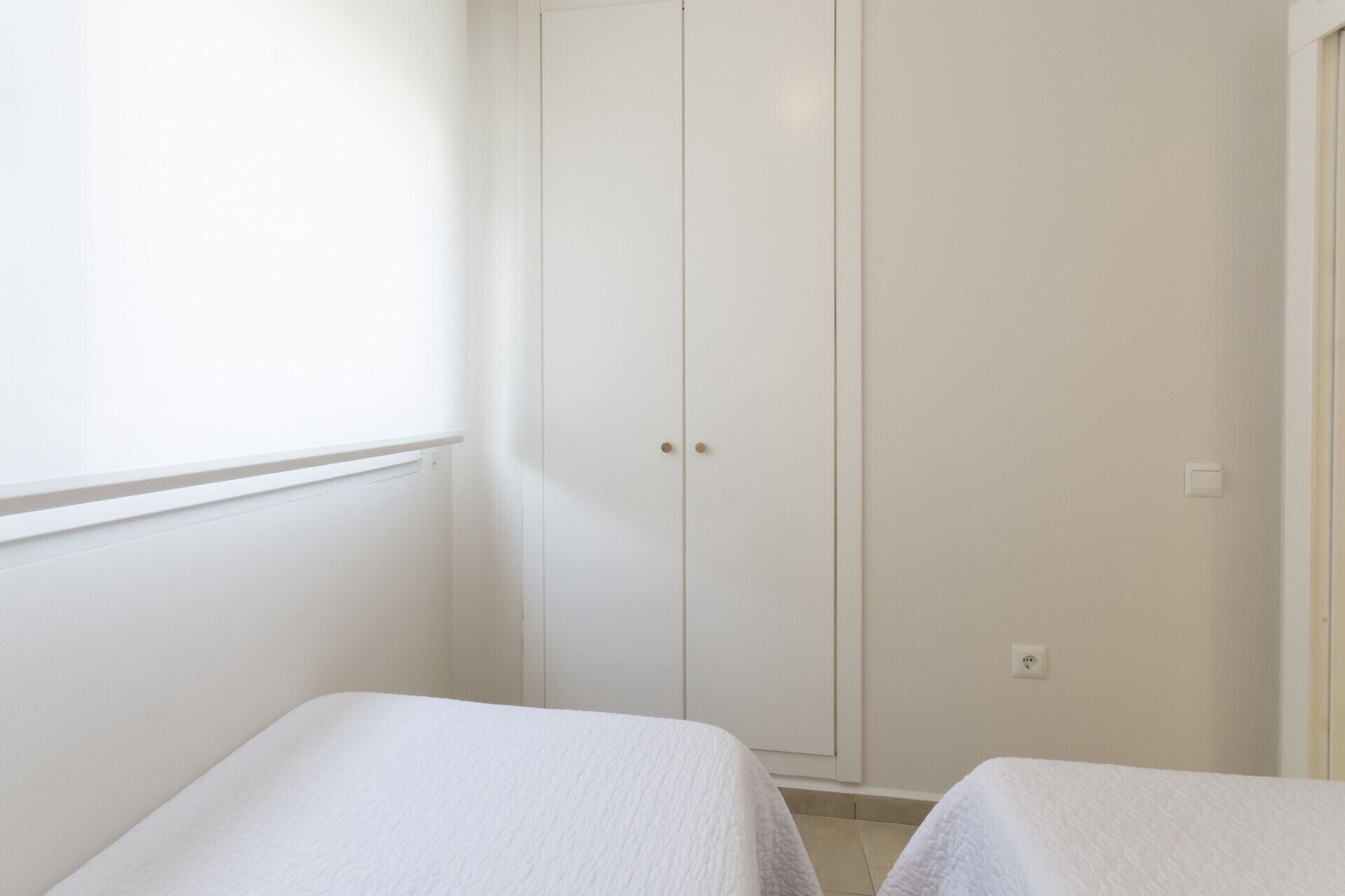 A small, white room with two single beds, a white wardrobe, and a window.