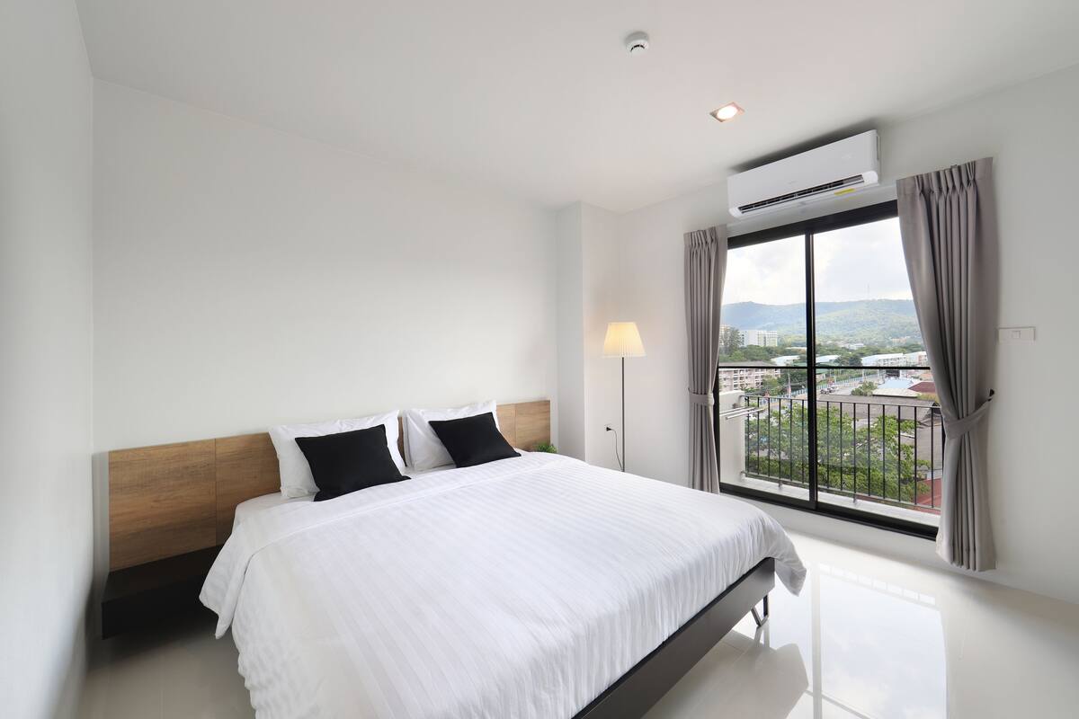 Una habitación de hotel moderna con una cama grande, cabecera de madera, vista de la ciudad a través de un ventanal amplio y un equipo de aire acondicionado.