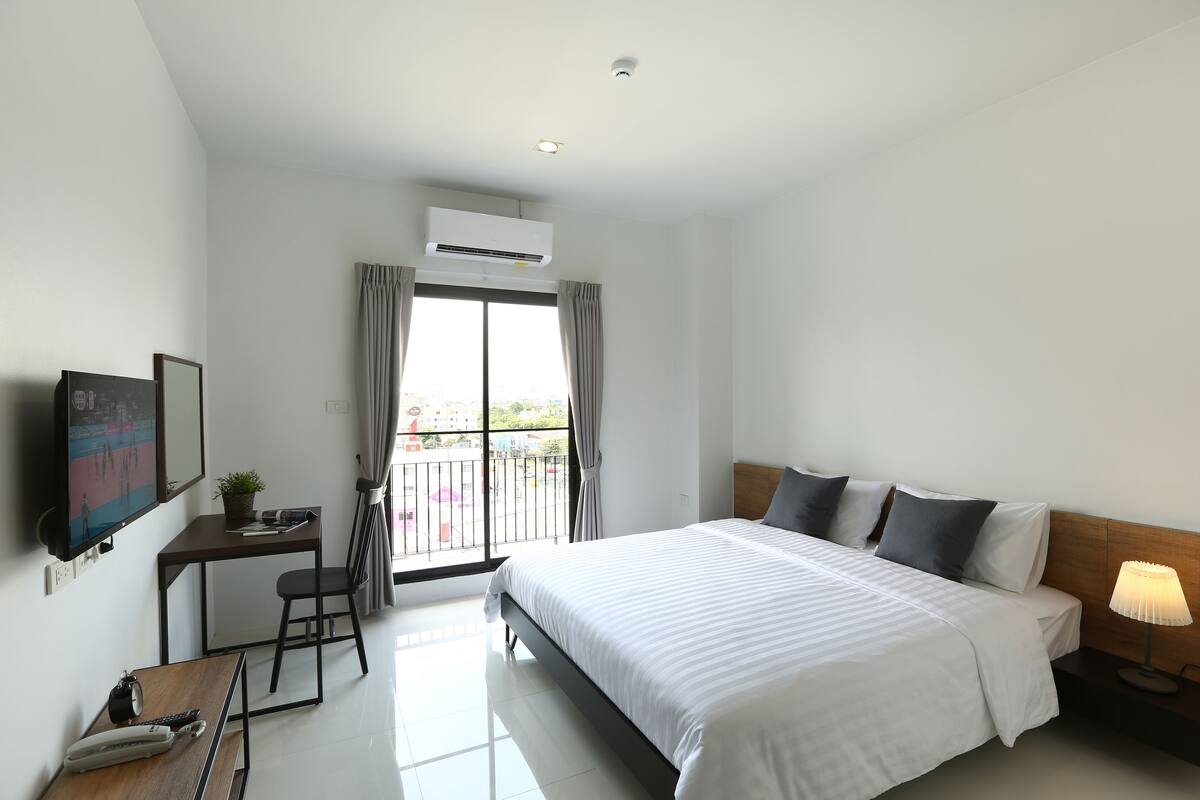 Habitación de hotel con cama, mesita de noche, escritorio, silla, televisor y balcón con cortinas.
