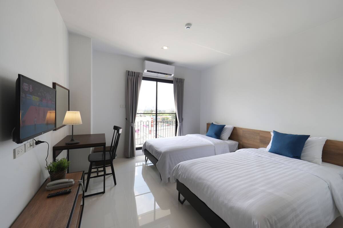 Habitación de hotel con dos camas, un escritorio con lámpara, televisión y balcón con cortinas.