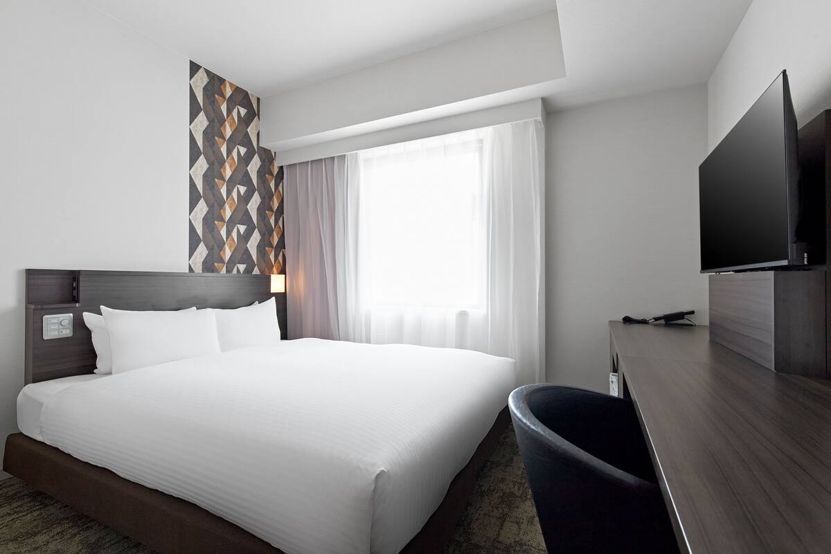 A hotel room with a large bed, a desk, a flat-screen TV, and a window with curtains.