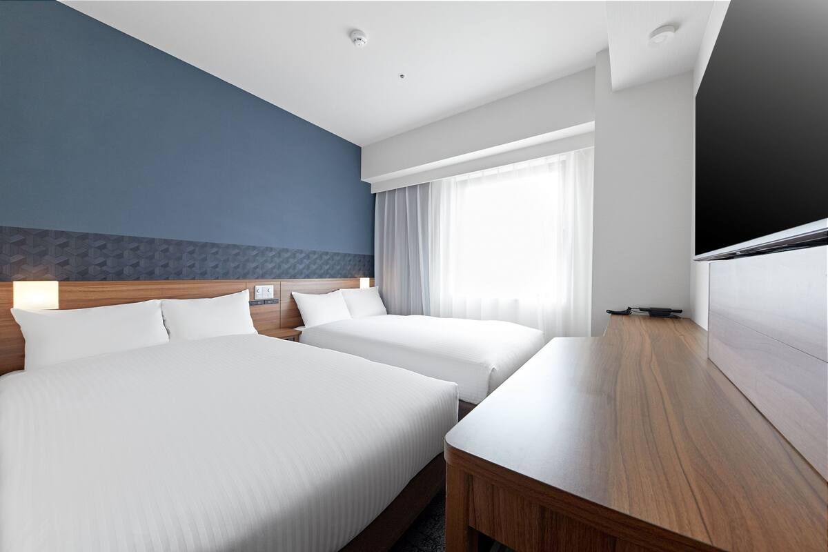 A hotel room with two beds, a wooden desk, a flat-screen TV, and a large window with curtains.