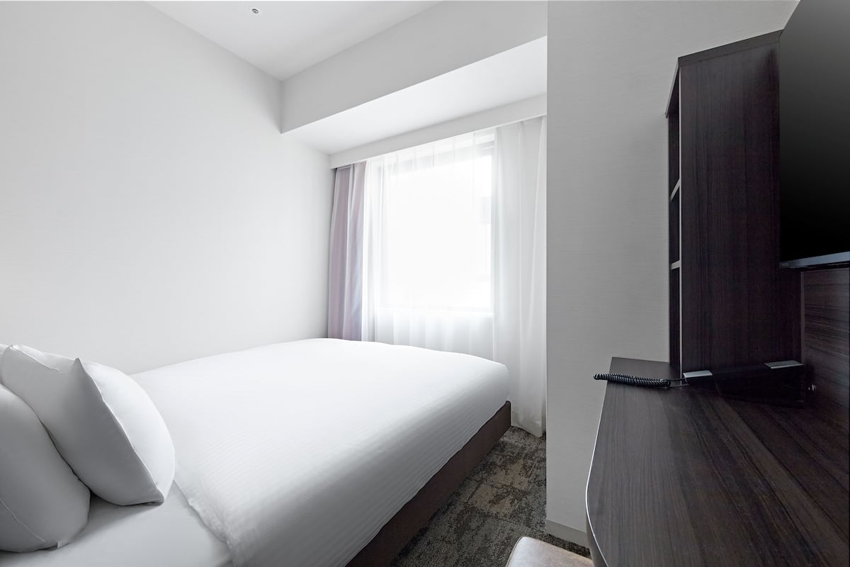 A hotel room with a single bed, a dark wooden dresser, a flat-screen TV, and a large window with sheer curtains.