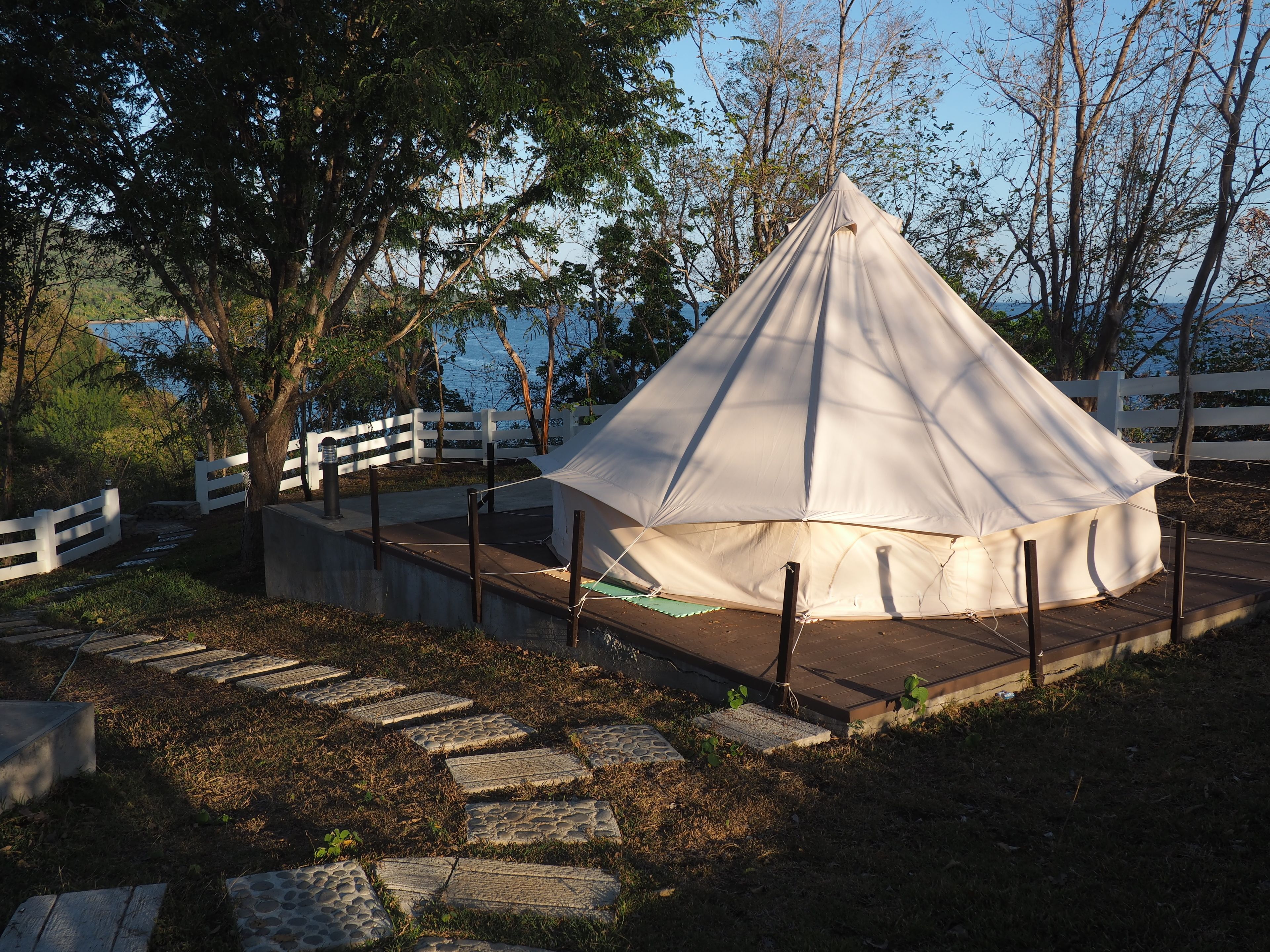 Uma tenda branca com telhado pontiagudo, cercada por uma cerca branca, sobre um caminho de pedra com grama e árvores ao fundo.