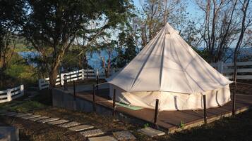 Uma tenda branca com telhado pontiagudo, cercada por uma cerca branca, sobre um caminho de pedra com grama e árvores ao fundo.