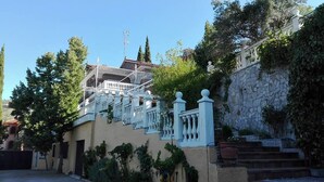 Ein Gebäude mit einer Treppe, die zu einem Balkon führt, umgeben von Grünpflanzen und einer Steinmauer.