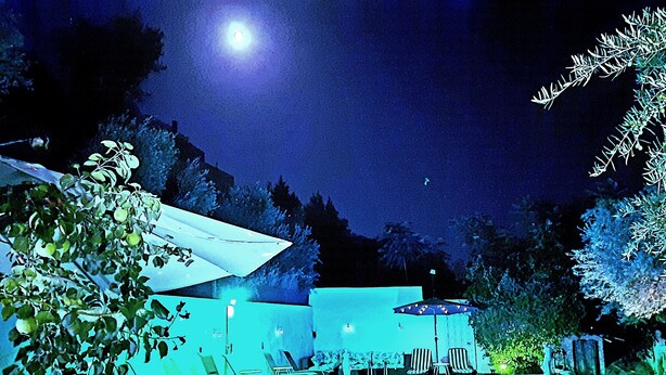 Ein Poolbereich im Freien bei Nacht mit Liegestühlen und Sonnenschirmen, umgeben von Bäumen und einem Berg im Hintergrund.