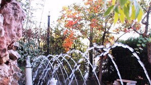 Ein Steinfbrunnen mit Wasser, das aus mehreren Bögen sprüht, umgeben von Pflanzen und einem Backsteinweg.