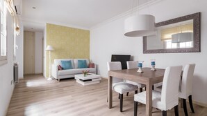 A modern living room with a dining area, featuring a wooden dining table, white upholstered chairs, a sofa with colorful cushions, a patterned wall, and a large mirror. - El Viso Smart / Smart Apartments Madrid (Madrid)