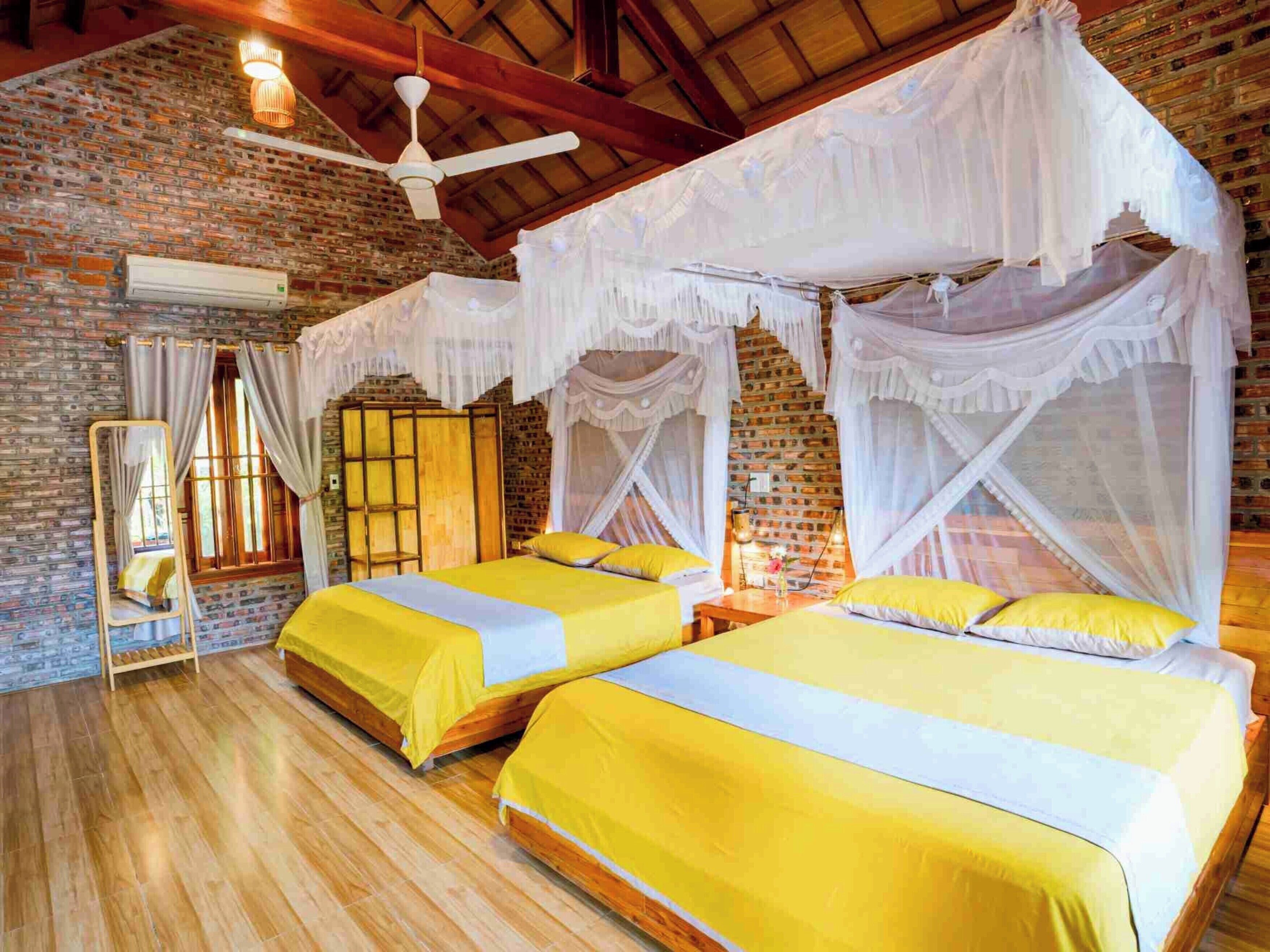 Una habitación con dos camas, ventilador de techo, pared de ladrillo y ventana con cortinas.