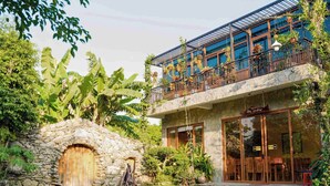 A two-story building with a stone archway entrance, a balcony with hanging plants, and a circular brick patio with a fire pit. - Ninh Binh Eco Garden (Tay Hoa Lu)