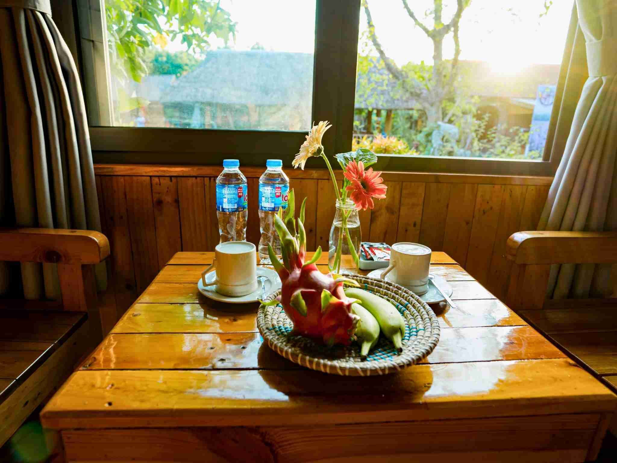 A wooden table with a vase of flowers, two water bottles, and two cups, likely for coffee or tea, in a room with wooden walls and a window overlooking greenery.