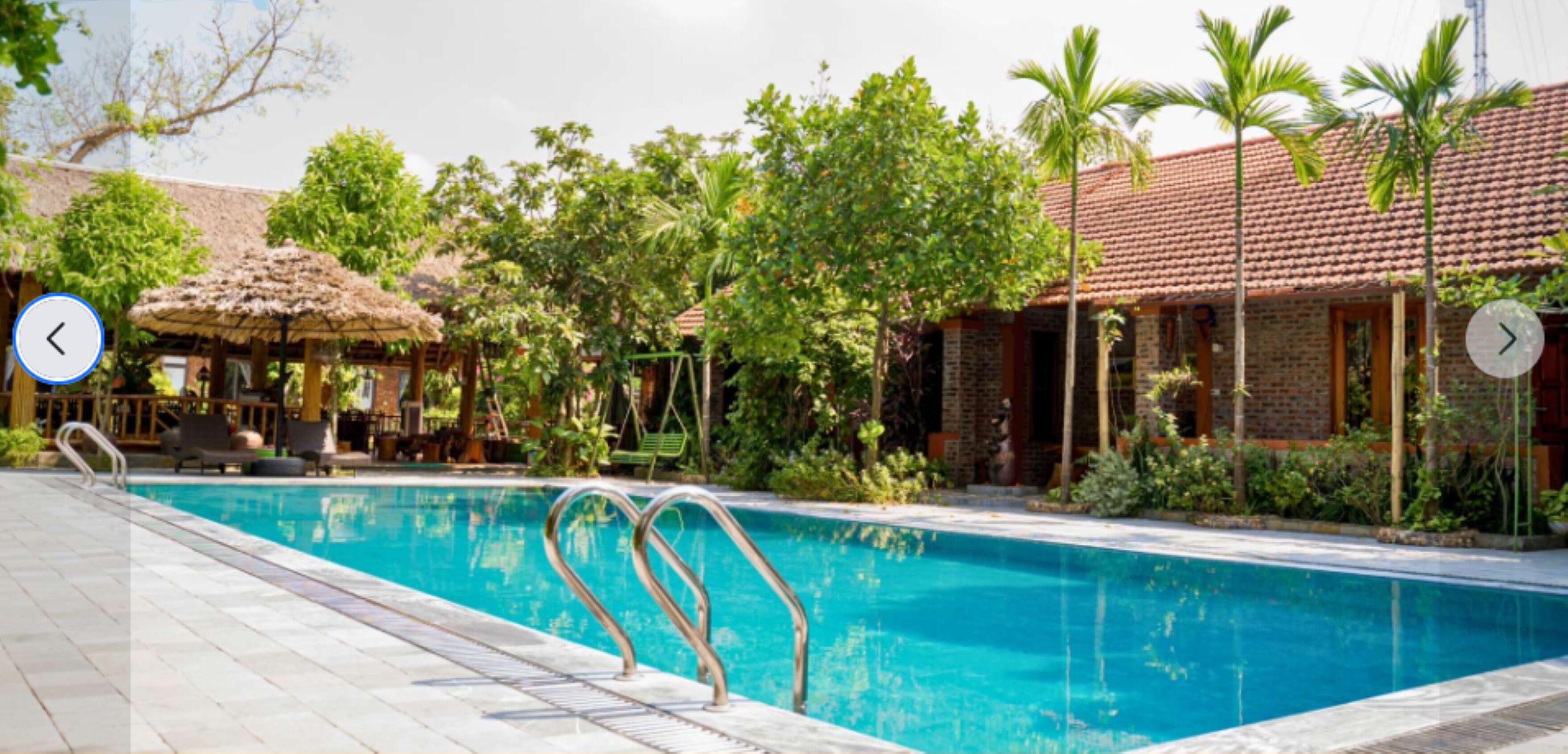 A swimming pool with a thatched-roof structure and a brick building in the background.