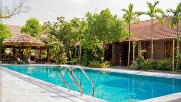 A swimming pool with a thatched-roof structure and a brick building in the background.