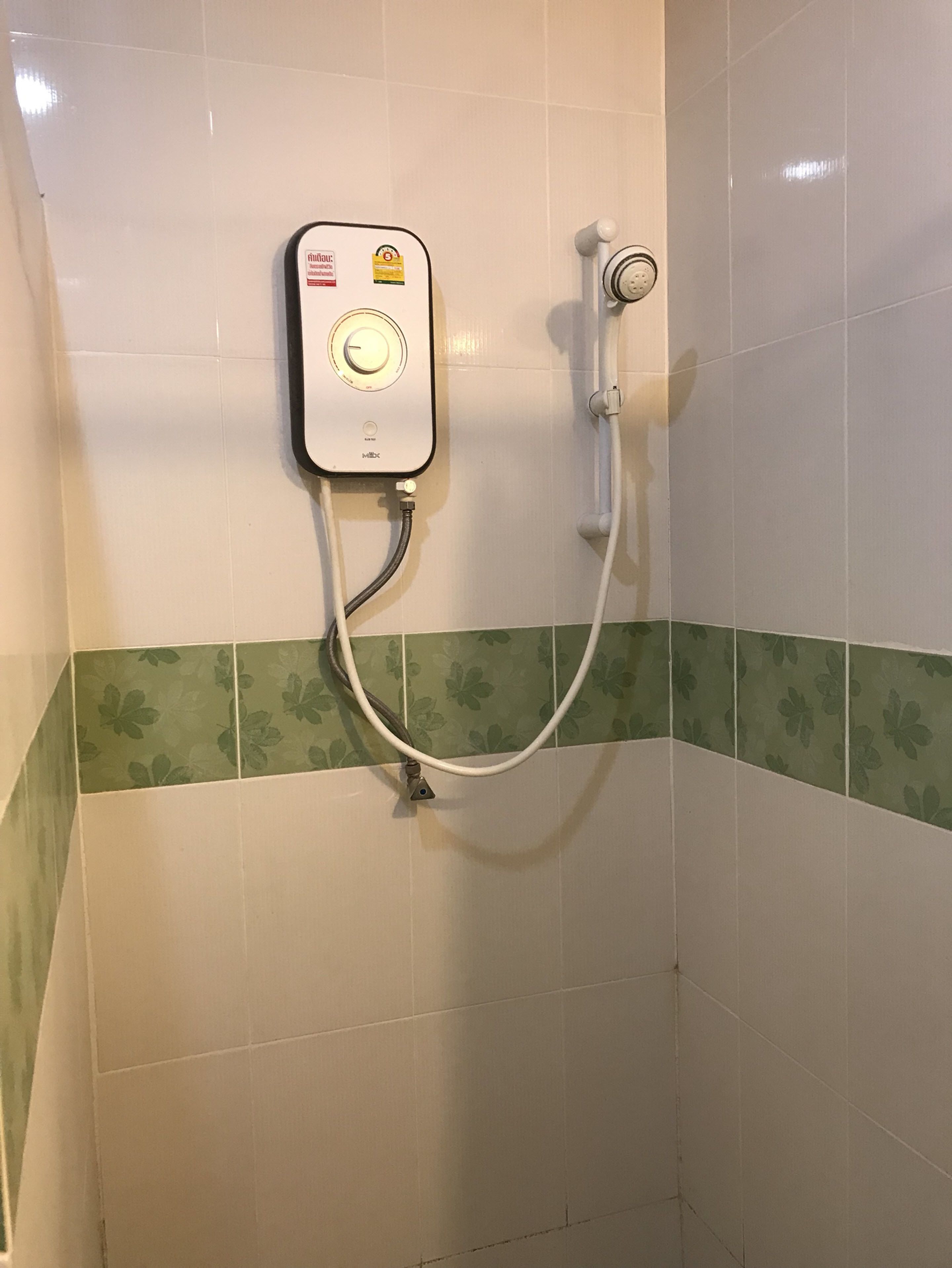 Una unidad de ducha con panel de control y cabezal de ducha de mano, instalada en una pared con azulejos florales verdes.