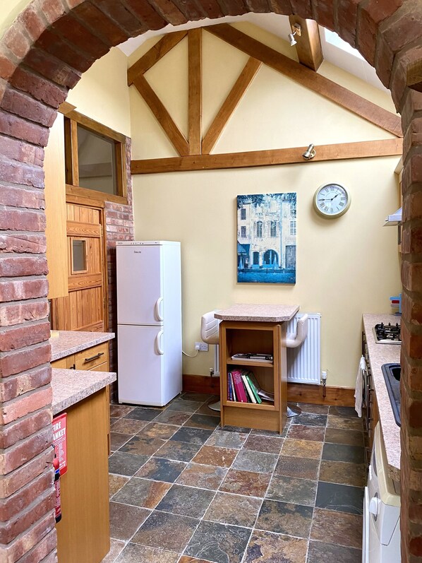 A compact kitchen with a refrigerator, a wooden cabinet, a shelf with books, a clock, and a tiled floor. - The Woodshed (Much Wenlock)