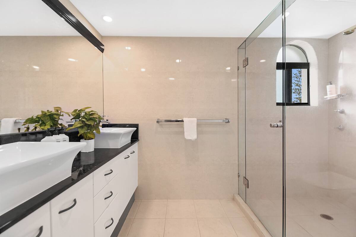 Un baño moderno con cabina de ducha de vidrio, un espejo grande y una encimera negra con armarios blancos.