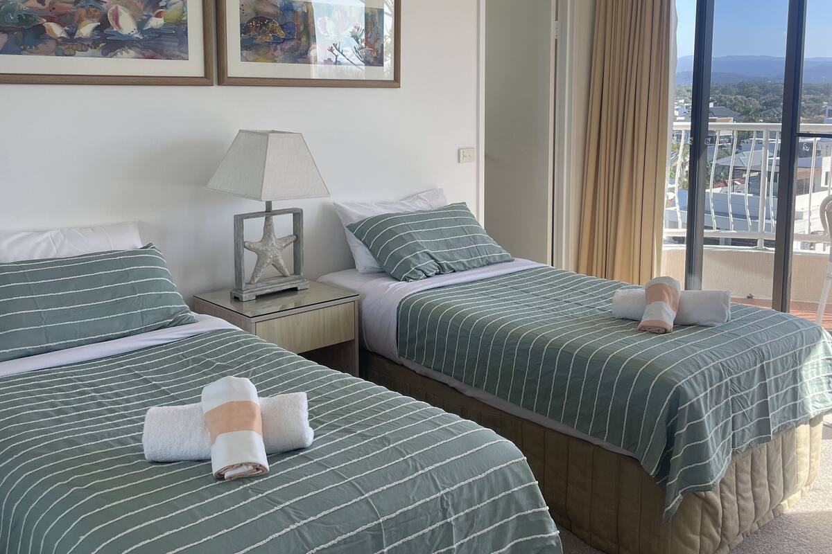 Habitación de hotel con dos camas, ventilador de techo, obras de arte en la pared y vistas de la ciudad a través de amplias ventanas.