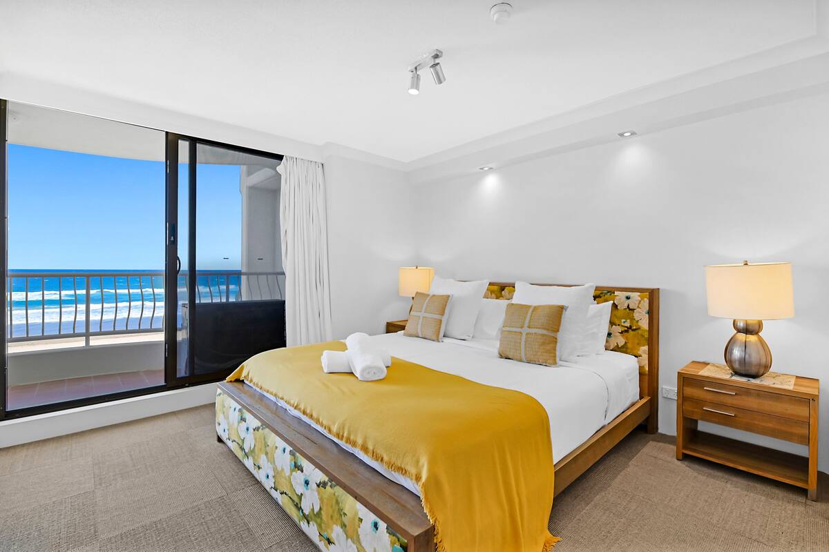 Habitación de hotel con una cama grande, dos lámparas de noche, una mesita de madera y vista al océano a través de una puerta corrediza de vidrio.