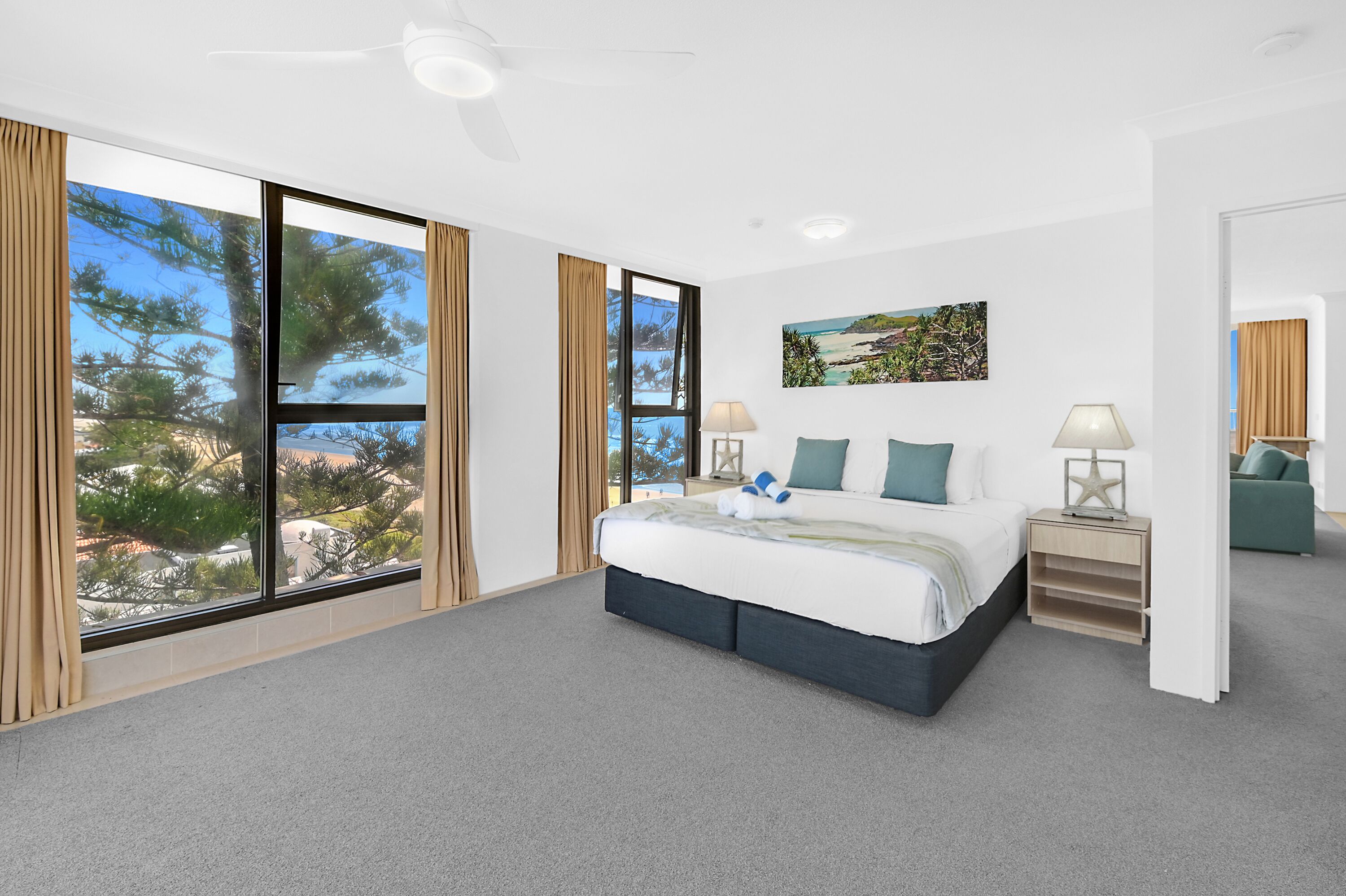 A bedroom with a large bed, a nightstand, a lamp, and a view of trees and buildings through large windows.