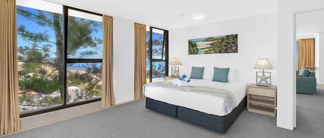 A bedroom with a large bed, a nightstand, a lamp, and a view of trees and buildings through large windows.