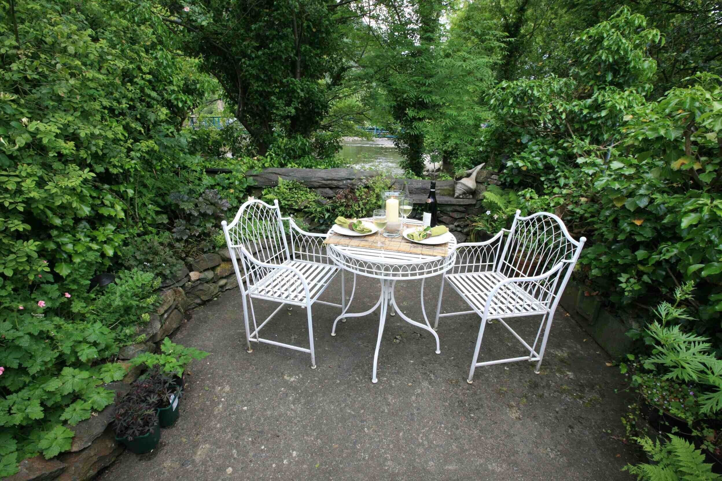 A garden patio with a round table and four white metal chairs, surrounded by lush greenery and a stone wall.