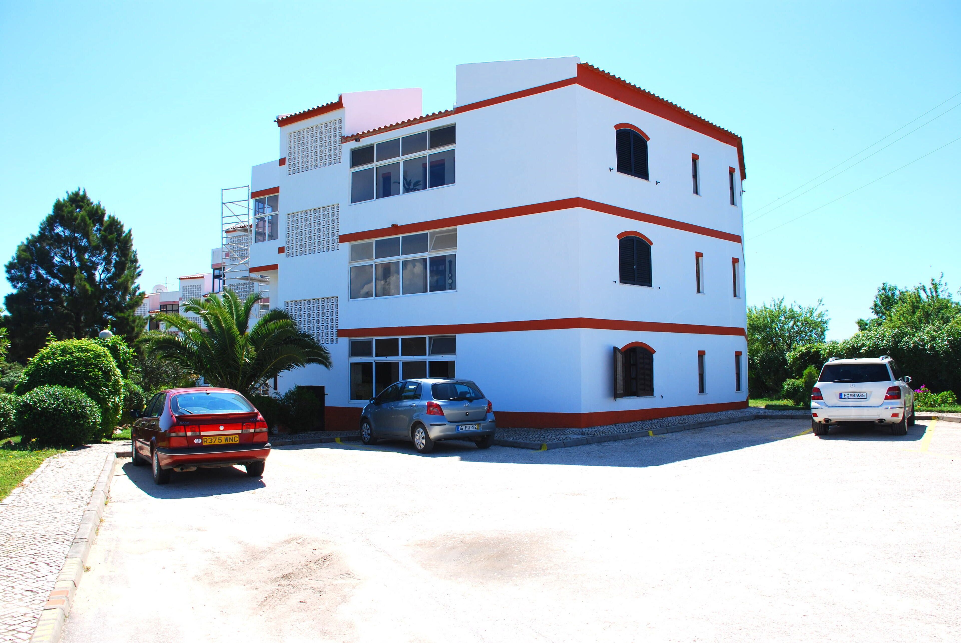 A multi-story building with a white facade and red accents, featuring a parking area with several cars.