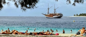 Una playa con gente tomando el sol y nadando, y un barco en el fondo.