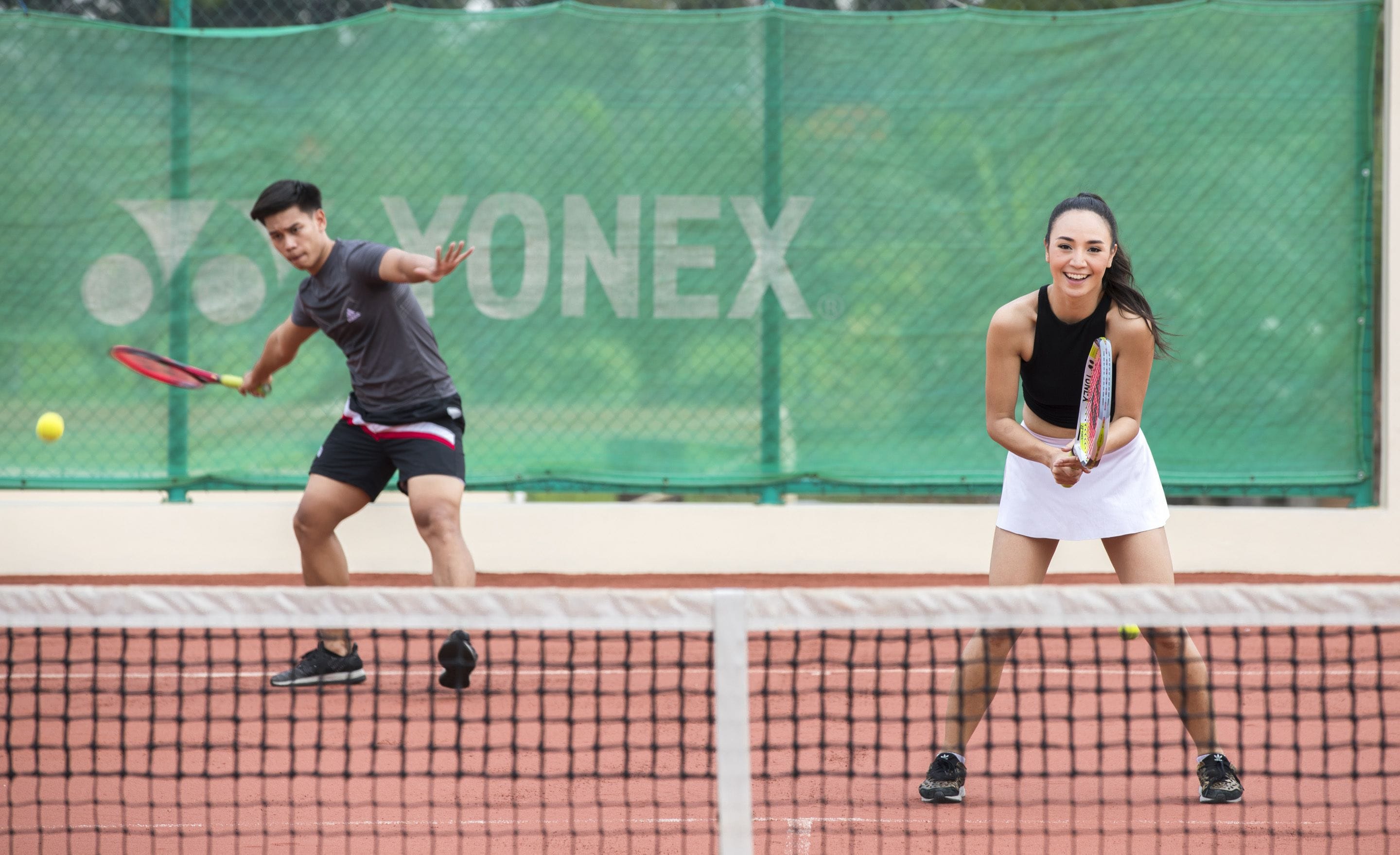 Una cancha de tenis con una red, un jugador con camiseta y pantalones cortos negros, y otra jugadora con falda blanca.