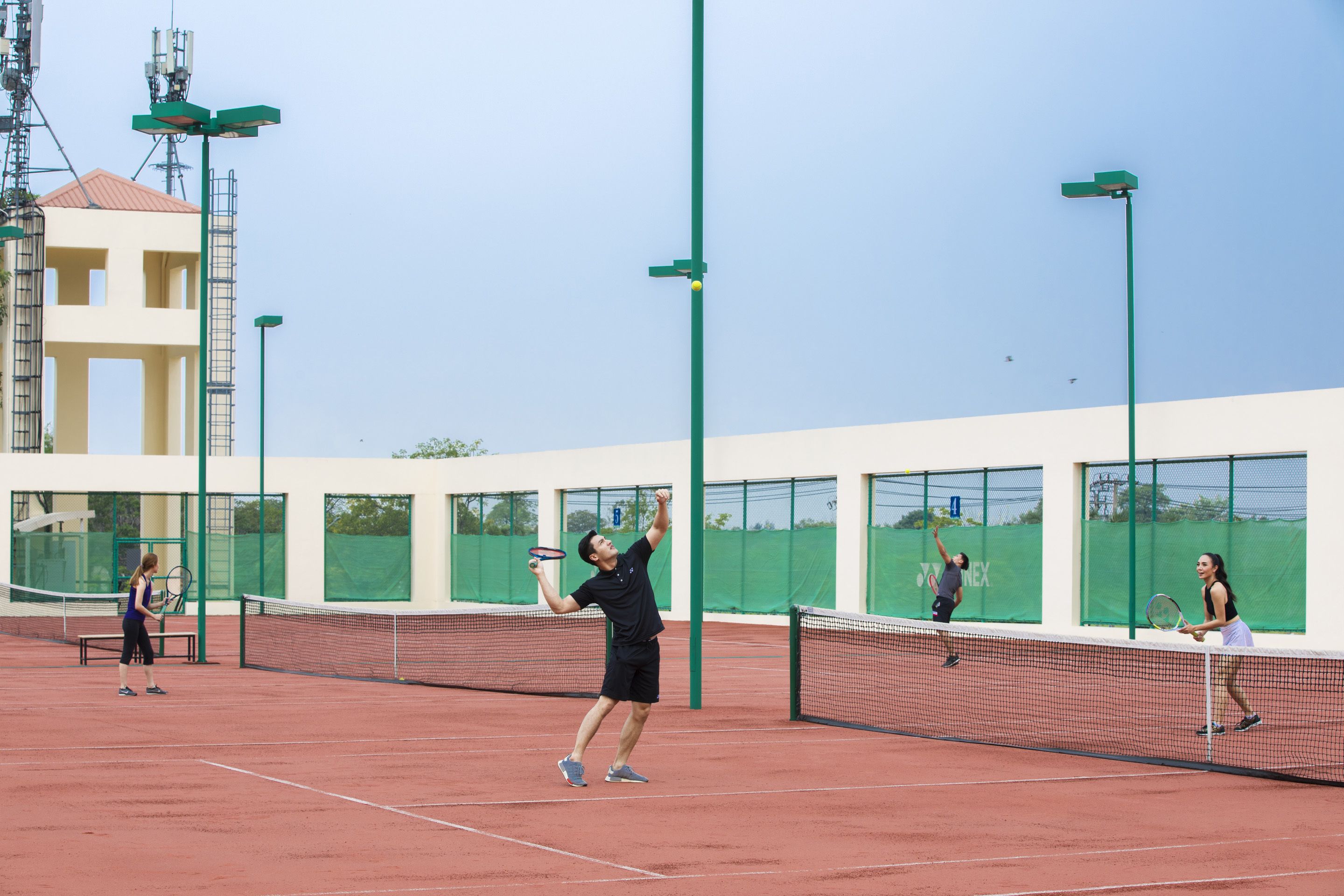 Un campo da tennis con rete, pali verdi illuminanti e un edificio sullo sfondo circondato da una recinzione verde.