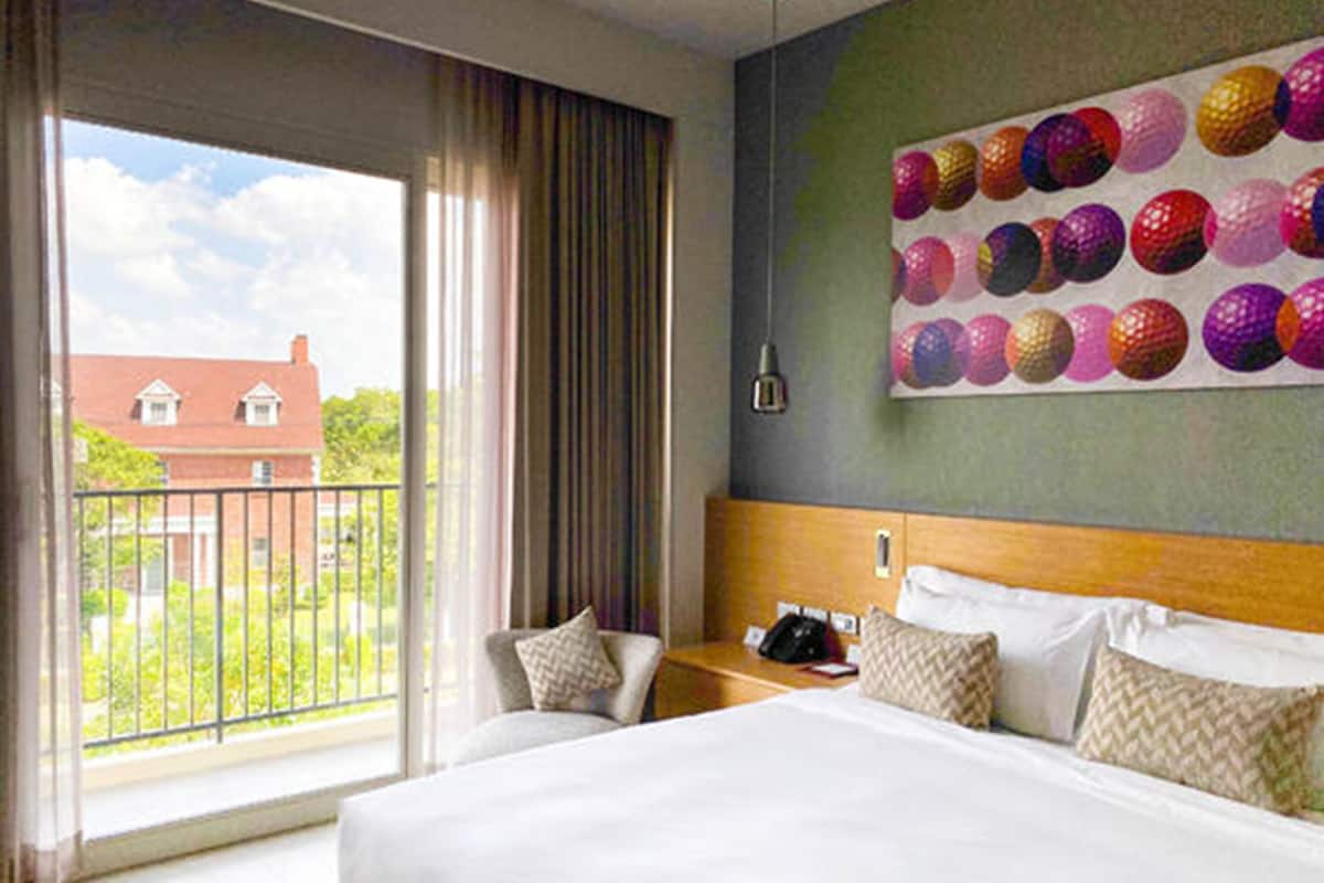 A hotel room with a large bed, a desk with a phone, and a view of a building through a window.