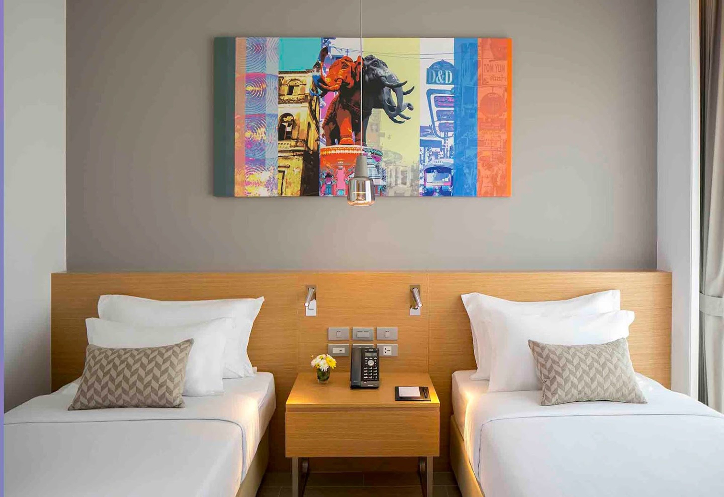 A hotel room with two beds, a nightstand with a phone and a vase of flowers, and a colorful cityscape painting above the beds.