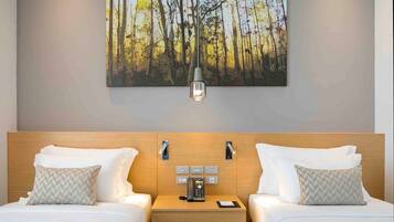A hotel room with two beds, a nightstand with a phone and a lamp, and a painting of a forest on the wall.